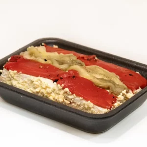 Un pastel de escalivada servido en un plato de cerámica, con su superficie dorada y acompañado de salsa romesco y pan tostado.