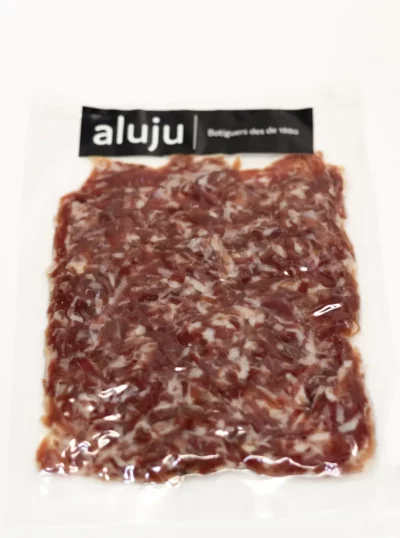 Virutas de jamón ibérico: sabor intenso en cada pequeño bocado Virutas de jamón ibérico cortadas a mano, con vetas de grasa y color intenso, servidas sobre tabla de madera.