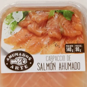 Carpaccio de salmón ahumado en finas lonchas, servido con hojas verdes y aliño de aceite y pimienta sobre plato blanco.