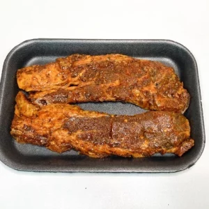 Churrasco adobado Aluju: sabor intenso para tus asados con carácter Dos tiras de churrasco adobado Aluju, con color rojizo y especias visibles, presentadas en bandeja negra, listas para cocinar a la parrilla, sartén o al horno.