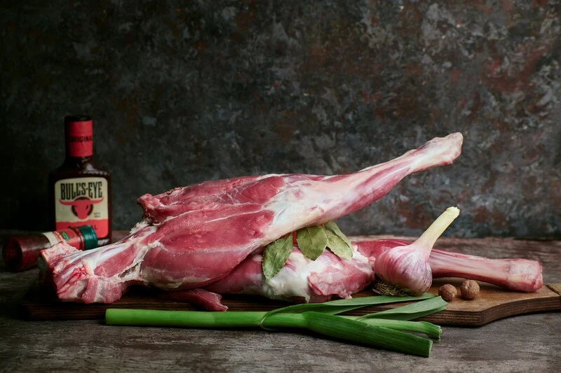 Pierna de cordero selección Aluju sobre tabla de madera, acompañada de ajo, laurel, cebollino y especias, lista para preparar al horno o a la brasa.