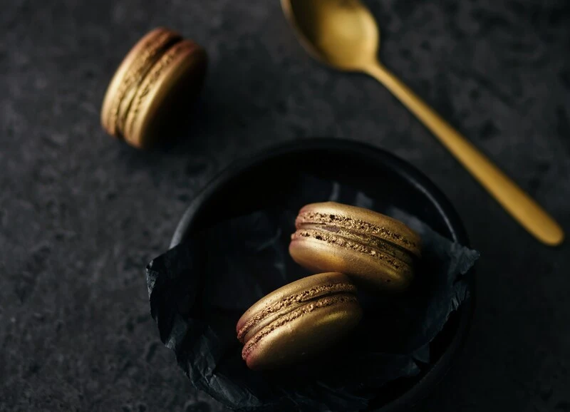 En Gourmet tres macarons dorados Aluju, dos en bol negro con papel decorativo y uno sobre superficie oscura, acompañados de cuchara dorada, que evocan lujo y alta repostería.