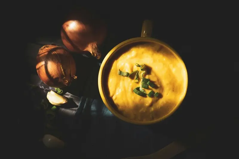 Bol de crema casera con cebollino picado, acompañado de cebollas y ajo fresco sobre fondo oscuro, evocando cocina tradicional y sabor reconfortante.