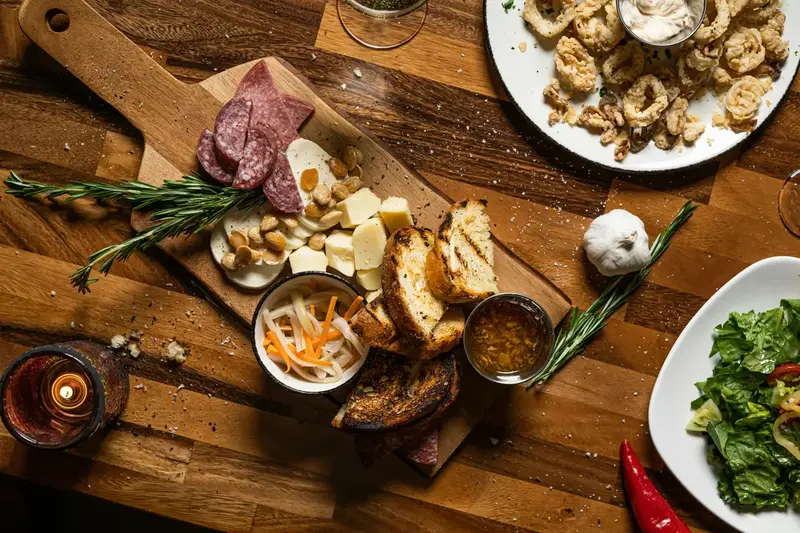 Selección de productos en otros, complementarios en Carns Aluju, incluyendo embutidos, quesos, pan tostado y aperitivos gourmet sobre una mesa de madera.
